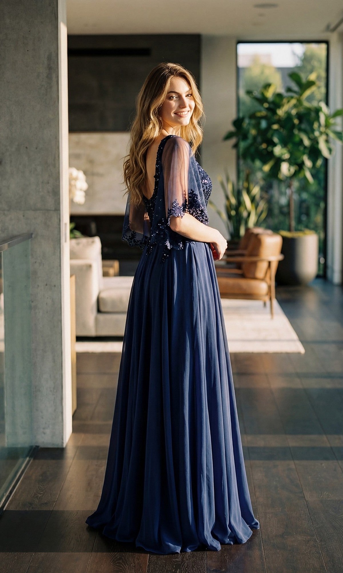 A woman stands smiling in a modern living room with large windows and plants, wearing the Juliet JTM22U long formal dress—featuring a navy blue flowy design with an embroidered sheer bodice and a graceful A-line, floor-length skirt.